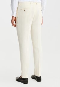 Kremowe lniane spodnie o slim fit, z tylnymi kieszeniami i detalem w postaci guzika. W połączeniu z czarnymi butami, widok z tyłu.