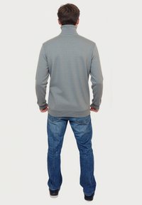 Grauer, strukturierten, hochgeschlossener Pullover mit langen Ärmeln, kombiniert mit blauen Jeans. Der Pullover hat gerippte Bündchen und einen gerippten Saum.