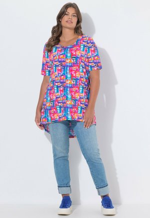 Młoda kobieta ubrana w kolorową, wzorzystą, oversize'ową bluzkę, jasnoniebieskie, podwinięte jeansy oraz jaskrawo niebieskie trampki, stojąca na białym tle.