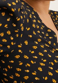 Blouse en tissu noir avec motif floral jaune, col en V et manches ballon froncées. Texture douce, motif réparti uniformément sur toute la surface.