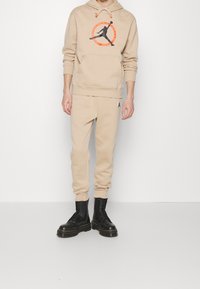 Felpa beige con logo grafico, pantaloni della tuta beige abbinati e stivali neri con lacci. L'outfit presenta un tessuto testurizzato e una vestibilità casual.