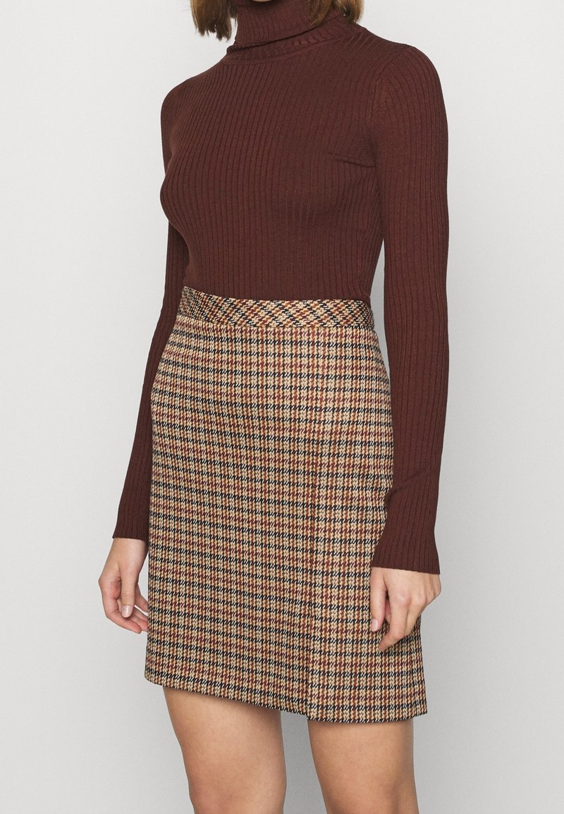 Femme portant un pull à col roulé côtelé marron et une jupe à carreaux mi-cuisse dans des tons beige, marron et noir sur un fond uni.