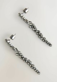 Orecchini in tonalità argento con un design a zigzag decorato con piccoli strass trasparenti, posizionati su uno sfondo liscio e neutro.