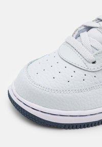 Närbild av tån på en vit texturerad lädersneaker, vita snören och en svart gummisula på vit bakgrund.