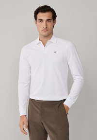 Mann trägt ein weißes Langarm-Polohemd mit kleinem Logo, braune Hose, steht vor einem einfarbigen hellgrauen Hintergrund, eine Hand in der Tasche.