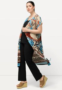 Cardigan in stile kimono multicolore con motivi floreali e geometrici, tessuto leggero, apertura frontale, indossato sopra una maglia nera e pantaloni neri.
