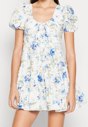 Kobieta ubrana w białą krótką sukienkę w niebieskie kwiaty, z bufkami, kwadratowym dekoltem i warstwową spódnicą.