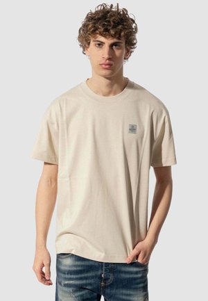 Giovane uomo con capelli ricci che indossa una T-shirt beige e jeans blu sbiaditi, in piedi davanti a uno sfondo grigio chiaro uniforme.