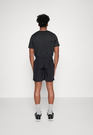 Schwarzes T-Shirt mit strukturiertem Design, getragen zu schwarzen Shorts. Das Outfit wird durch sportliche Schuhe und graue Socken ergänzt. Rückansicht.