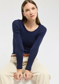 Jeune femme avec de longs cheveux bruns portant un haut à manches longues bleu marine et un pantalon beige, assise les mains jointes entre ses jambes.