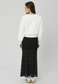 Pull blanc duveteux à ourlet arrondi associé à une jupe longue noire en dentelle. Le mannequin porte des baskets blanches, affichant un look à la fois décontracté et élégant.