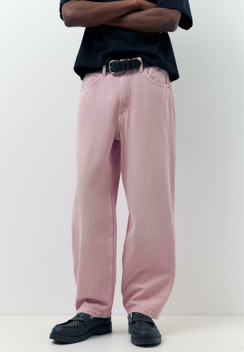 Homme portant un pantalon ample rose pâle avec une ceinture noire, des chaussures noires et une chemise foncée, bras croisés devant un fond blanc uni.