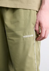 Olivengrüne Adidas-Hose mit glatter Textur, weißen Logodetails und einem kontrastierenden weißen Seitenstreifen. Nahaufnahme eines Hand, das den Stoff hält.