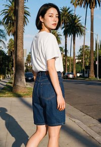 Tânără cu păr negru scurt, purtând tricou alb și pantaloni scurți din denim, mergând pe o stradă însorită plină de palmieri.