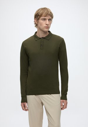 Bărbat cu păr deschis, purtând o cămașă polo verde închis cu mânecă lungă și pantaloni bej, stând pe un fundal alb simplu.