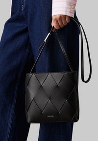 Borsa a mano in pelle intrecciata nera con forma rettangolare e tracolla regolabile. Presenta un dettaglio con logo nella parte inferiore. Texture liscia.