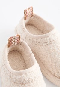 Paire de chaussons d'intérieur doux, de couleur crème, texturés, avec des bords rembourrés et de petites languettes en tissu tissé dans des tons rose, blanc et beige.