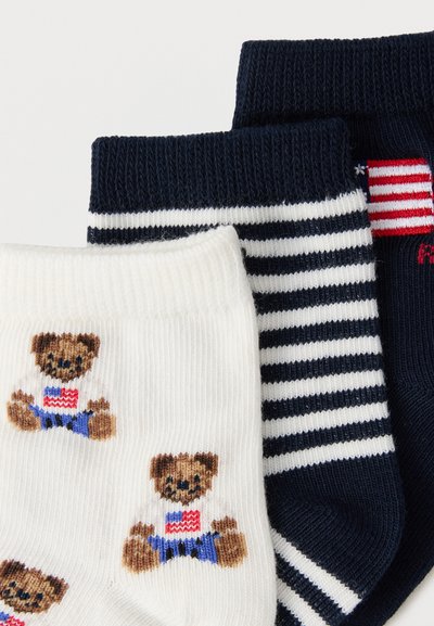 Τρία ζευγάρια διπλωμένες κάλτσες: ένα λευκό με αρκουδάκια που φορούν πουκάμισα με τη σημαία των ΗΠΑ, ένα μπλε σκούρο με λευκές ρίγες, ένα μονόχρωμο μπλε σκούρο με ένα μικρό πατς σημαίας.