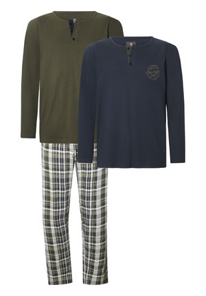 Bearded man wearing an olive green long-sleeve shirt and white, black, and green plaid pajama pants, standing barefoot against a plain background.