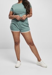 Kvinna med långt hår iklädd teal-färgad t-shirt och matchande shorts med vit kant, stående med benen isär och händerna knäppta.