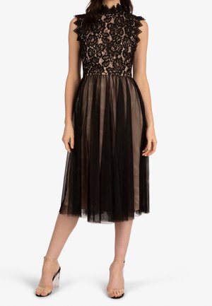 Robe noire sans manches en dentelle avec un corsage à motif floral et une jupe midi en tulle transparent, portée avec des sandales à bride cheville nude à talons transparents.