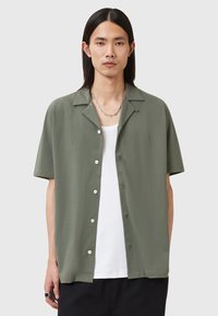 Korte ermer, krage skjorte i olivengrønn stoff; har knappelukking foran, subtilt merke, og en avslappet passform over en hvit tanktop.
