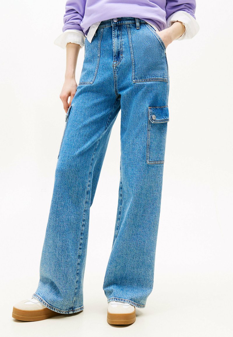 Jeans anchos azules en material de mezclilla con cintura alta, bolsillos frontales y un bolsillo lateral tipo cargo. Acabados con un dobladillo casual y combinados con zapatillas.