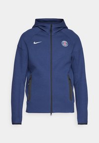 Veste à capuche bleu marine avec fermeture éclair sur le devant, poches latérales zippées, logo Nike blanc sur la poitrine droite et emblème du Paris Saint-Germain sur la poitrine gauche.