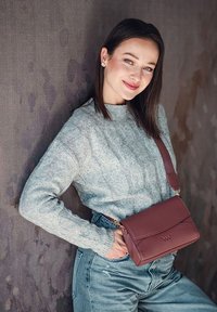 Braune Leder-Umhängetasche mit Klappenverschluss, geprägtem Logo und einem verstellbaren Tragegurt, kombiniert mit einem hellgrauen strukturierten Pullover und Jeans.