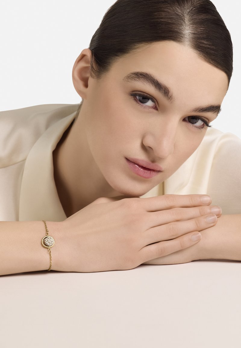 Kuldne käevõru, millel on ümmargune ripats tekstureeritud pinnaga. Kantud heledanahalisel käel, neutraalse taustaga.