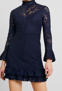 Robe en dentelle marine avec des manches longues, un col montant, un ourlet volé, un motif floral, un tissu texturé et des poignets de manches évasés.