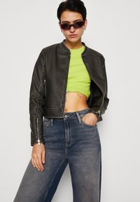 Veste bomber en cuir noir avec une fermeture éclair, des coutures contrastées et deux poches latérales zippées. Associée à un crop top vert et un jean bleu.