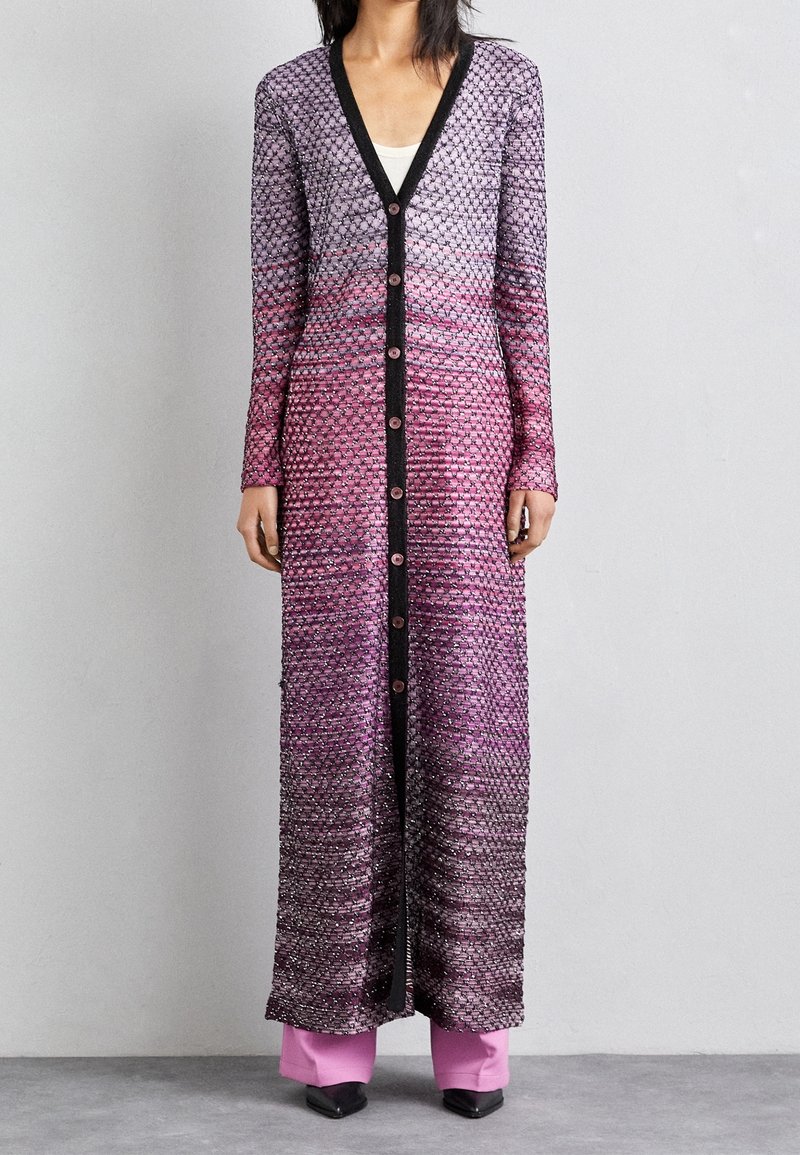 Femme portant un long cardigan violet-rose boutonné avec des bordures noires, un pantalon rose à jambes larges et des chaussures noires pointues, debout contre un mur uni.