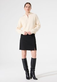 Suéter de canalé color crema con cuello de cremallera, combinado con una falda mini negra con una abertura lateral y botas negras de tacón alto hasta la rodilla.