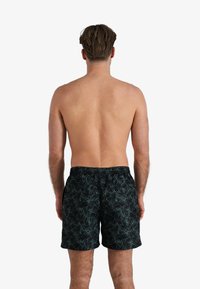 Zwarte zwembroek met een groen palmbladenpatroon, elastische tailleband en een ontwerp dat boven de knie valt, zichtbaar vanaf de achterkant.