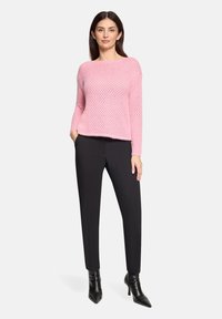 Femme portant un pull en maille rose, un pantalon noir ajusté et des bottines noires à bout pointu, debout avec une main dans la poche.