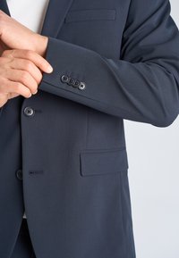 Person som justerer mansjetten på en mørk marineblå dressjakke med fire knapper og en lomme foran.