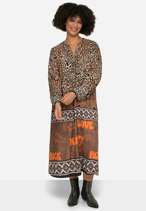 Leopardenmuster-Kleid in erdigen Tönen mit Kragen, langen Ärmeln und dekorativem orangem Text. Schwarze, mit Nieten besetzte Stiefeletten runden das Outfit ab.