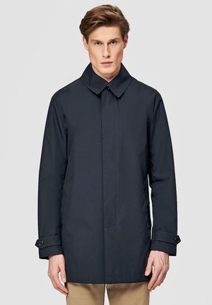 Młody mężczyzna o jasnych włosach, ubrany w zapinany na guziki ciemnoniebieski płaszcz i beżowe spodnie na jednolitym jasnoszarym tle.