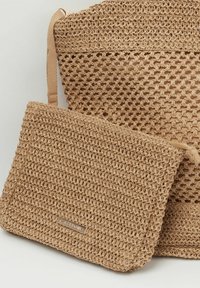 Bolso de paja tejido en color beige, con una superficie texturizada y un bolso plano estilo sobre extraíble con un sutil acento de logotipo.
