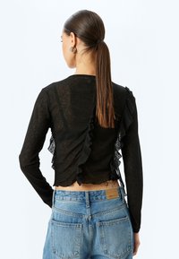 Schwarzes Langarmoberteil mit Spitzenruffdetails auf der Rückseite, kombiniert mit hellblauen, hochgeschnittenen Jeans mit Gesäßtaschen.