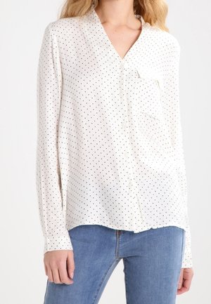 White long-sleeve blouse with small black polka dots, front chest pocket, buttoned cuffs, worn with blue denim jeans on a standing person.