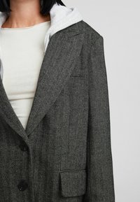 Blazer grigio a spina di pesce con un design strutturato, rever a punta, tre bottoni neri e una tasca frontale, indossato sopra un top bianco con cappuccio.