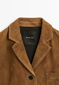 Giacca in suede marrone con colletto a punte, chiusura con un solo bottone e taschino sul petto. L'etichetta interna mostra il marchio e le informazioni per la cura.