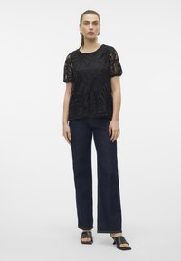 Sort blonde top med korte ærmer, der har detaljerede blomsterudskæringer. Kombineret med mørkeblå bredbenede jeans og sorte åben-tå sko.
