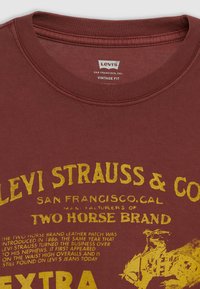 Bordeaux T-shirt met gele vintage-stijl grafische tekst. Katoenen stof met een ronde hals en korte mouwen. Opvallende illustratie van het "Two Horse Brand".