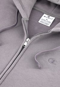 Sivá mikina na zips vyrobená z mäkkého materiálu, so zipsom na prednej strane, kapucňou so šnúrkou a vyšitým logom na ľavej hrudi. Viditeľná etiketa s pokynmi na údržbu.
