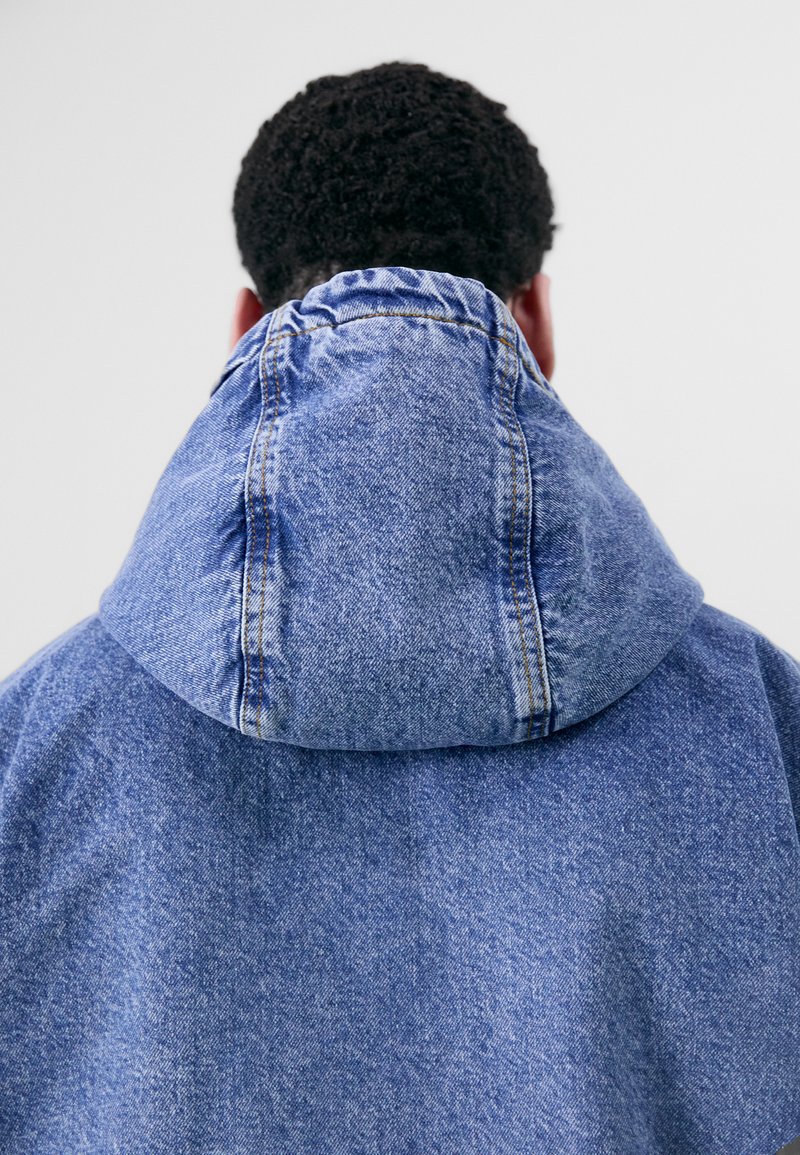 Giacca di jeans con un ampio cappuccio, caratterizzata da un lavaggio blu, cuciture visibili e una superficie liscia. La parte posteriore è piegata al colletto.