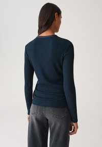 Frau mit langen dunklen Haaren, die ein enges, dunkelblaues, geripptes Langarm-Oberteil und lockere graue Jeans trägt, von hinten gezeigt.