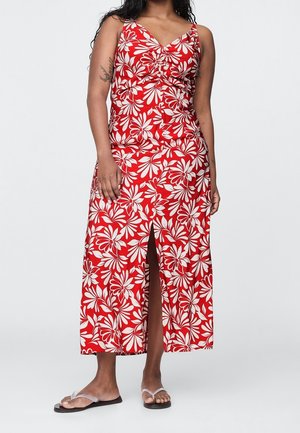 Femme portant une robe maxi rouge sans manches avec un motif floral blanc et une fente à l'avant, associée à des sandales plates à brides transparentes.